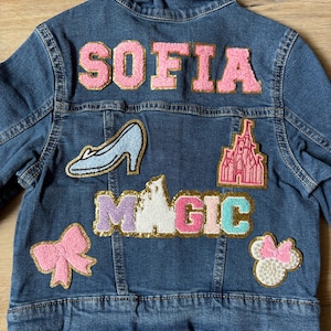May include: A blue denim jacket with various colorful patches, including a pink bow, a blue glass slipper, a pink castle, a multi-colored "MAGIC" word, and a pink and white Minnie Mouse head. The name "SOFIA" is written in pink letters with gold outlines on the back of the jacket.