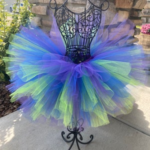 Puede incluir: Una falda de tut&uacute; de tul de varias capas en tonos de morado, azul y verde. La falda se muestra en un soporte de metal negro.