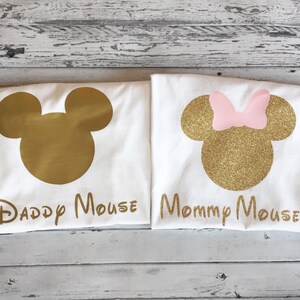 May include: Two white t-shirts with gold glitter Mickey Mouse silhouettes. The left shirt says "Daddy Mouse" and the right shirt says "Mommy Mouse".