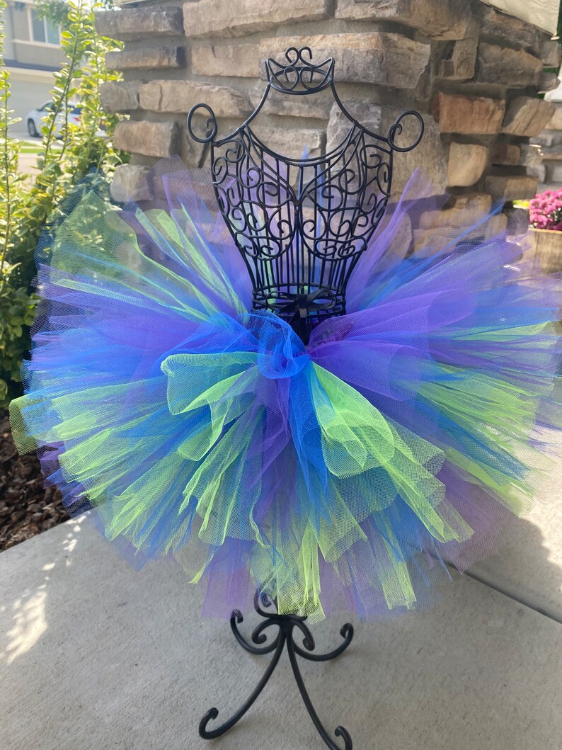 Puede incluir: Una falda de tut&uacute; de tul de varias capas en tonos de morado, azul y verde. La falda se muestra en un maniqu&iacute; de metal negro.