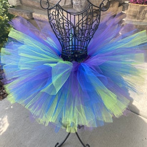 Puede incluir: Una falda de tut&uacute; de tul multicolor con tonos de azul, morado y verde. La falda se muestra en un soporte de metal negro.