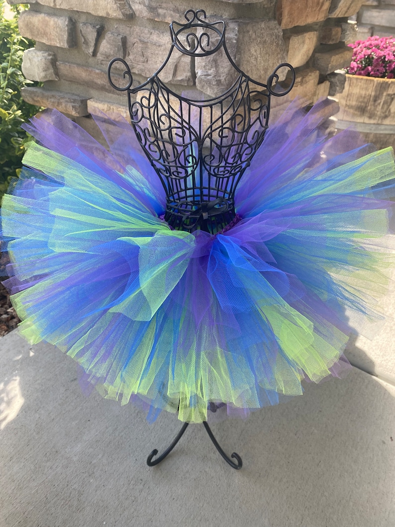 Puede incluir: Una falda de tut&uacute; de tul multicolor con tonos de azul, morado y verde. La falda se muestra en un soporte de metal negro.