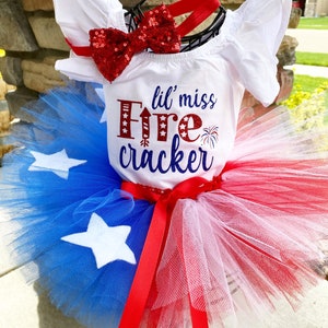 K&ouml;nnte beinhalten: Ein wei&szlig;er Baby-Strampler mit dem Text "lil' miss Firecracker" in roten und blauen Buchstaben. Der Strampler wird mit einer roten Pailletten-Schleife und einem roten, wei&szlig;en und blauen T&uuml;ll-Tutu kombiniert.