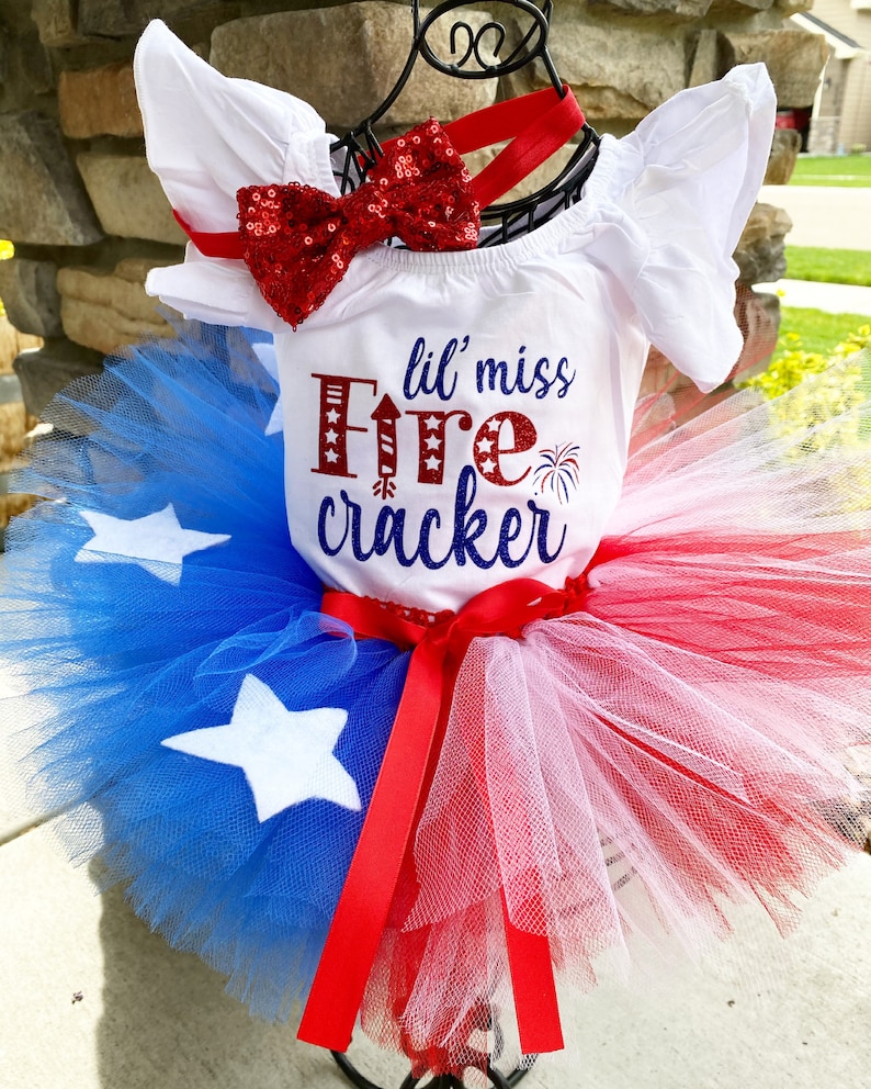 K&ouml;nnte beinhalten: Ein wei&szlig;er Baby-Strampler mit dem Text "lil' miss Firecracker" in roten und blauen Buchstaben. Der Strampler wird mit einer roten Pailletten-Schleife und einem roten, wei&szlig;en und blauen T&uuml;ll-Tutu kombiniert.