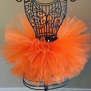 May include: A bright orange tulle tutu skirt on a black mannequin. The skirt is full and fluffy, with a soft, delicate texture.