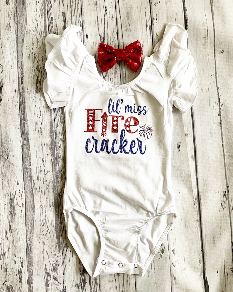 K&ouml;nnte beinhalten: Wei&szlig;er Body mit rotem und blauem Glitzertext, der "lil' miss firecracker" lautet. Ein rotes Paillettenband ist enthalten.