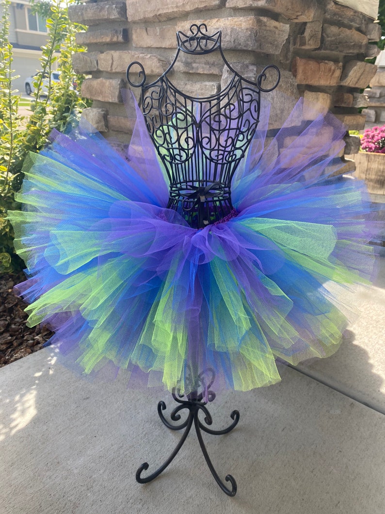 Puede incluir: Una falda de tut&uacute; de tul de varias capas en tonos de morado, azul y verde. La falda se muestra en un soporte de metal negro.