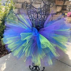 Puede incluir: Una falda de tut&uacute; de tul de varias capas en tonos de morado, azul y verde. La falda se muestra en un maniqu&iacute; de metal negro.