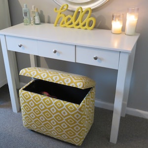 May include: White vanity table with two drawers and a yellow "hello" sign on top. A yellow and white patterned storage ottoman sits in front of the table.
