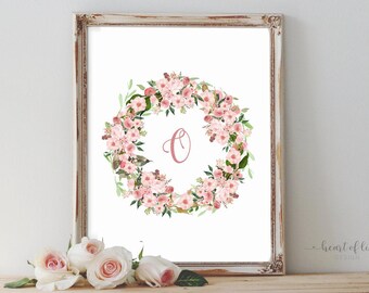 May include: A framed print with a floral wreath design. The wreath is made of pink flowers and green leaves, encircling a pink letter "O". The frame has a distressed, off-white finish. Several roses are in the foreground, adding to the floral theme.