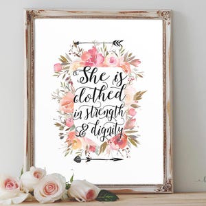 May include: Framed art print with a floral wreath design. The print features the quote "She is clothed in strength & dignity" in elegant script. The frame is a distressed white with a decorative edge. Soft pink and peach flowers surround the text.