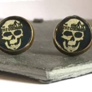 May include: A pair of gold-tone skull earrings with a black background and the text "The Goonies" in white.
