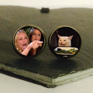 Könnte beinhalten: Ein Paar runde, silberne Manschettenknöpfe mit Bildern von beliebten Internet-Memes. Ein Manschettenknopf zeigt eine Frau, die ein Katze anschreit, und der andere Manschettenknopf zeigt eine Katze, die auf einen Teller mit Essen schaut.