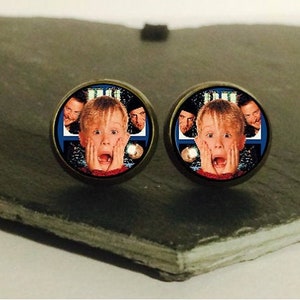 Peut inclure: Une paire de boucles d'oreilles rondes argentées avec une image d'un jeune garçon avec une expression surprise. Le garçon est entouré d'autres personnes sur l'image. Les boucles d'oreilles sont sur une surface en forme de cœur gris foncé.