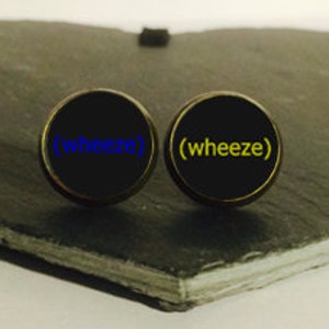 May include: Two black round buttons with a gold rim. The left button has the word "wheeze" in blue text. The right button has the word "wheeze" in yellow text.