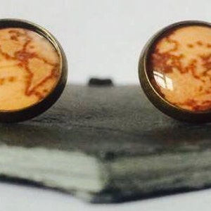 Puede incluir: Un par de pendientes de botón dorados con un diseño de mapa del mundo. El mapa está impreso sobre un fondo naranja claro con líneas marrones.