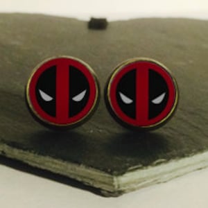 Peut inclure: Une paire de boucles d'oreilles rondes en argent avec un motif de logo Deadpool rouge et noir.