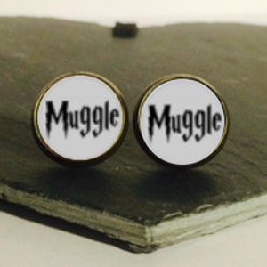 Puede incluir: Dos pendientes redondos, blancos, de estilo botón con la palabra "Muggle" impresa en negro en cada pendiente.