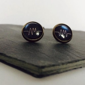 Könnte beinhalten: Ein Paar goldfarbene Ohrstecker mit schwarzem Hintergrund und einem weißen Wellenform-Design.