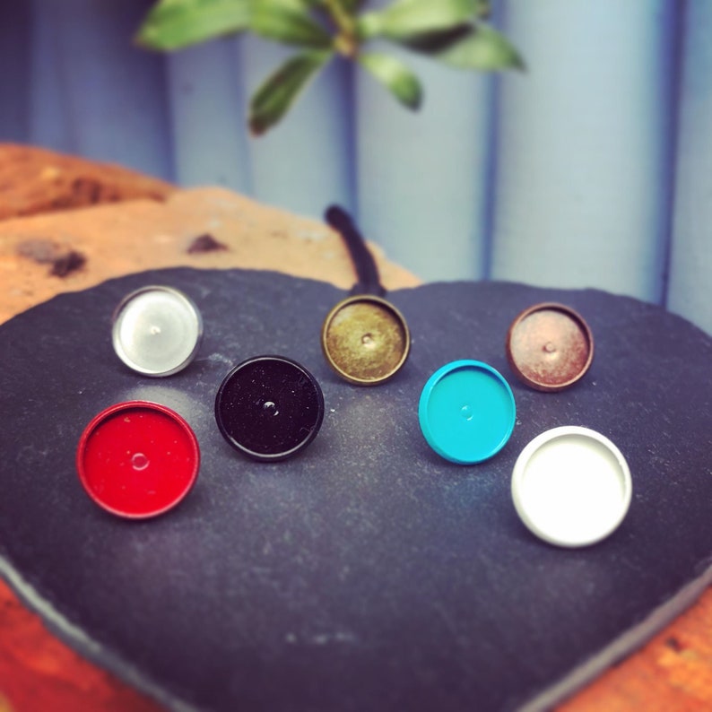 May include: Assortment of eight round, flat, metal earring blanks in various colors, including black, white, red, blue, gold, and silver. The blanks are arranged on a black surface.