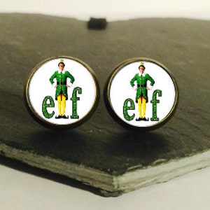 Op de afbeelding: Een paar ronde, witte, knoopvormige oorbellen met een gouden rand. De oorbellen hebben een cartoonafbeelding van Buddy the Elf uit de film Elf, die een groen pak en een groene hoed draagt. De tekst "elf" is in groene letters onder de afbeelding gedrukt.