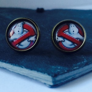 May include: A pair of silver stud earrings with a round, clear glass design featuring the Ghostbusters logo. The logo is a red no-ghost symbol with a white ghost inside.