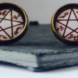 Può includere: Un paio di orecchini con un design a pentagramma e l'iscrizione "veneris" in rosso su sfondo bianco. Gli orecchini sono in metallo e hanno una finitura bronzo.