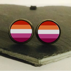 Peut inclure: Une paire de boucles d'oreilles clous avec un motif de drapeau de la fierté lesbienne. Les boucles d'oreilles sont rondes et présentent un motif de drapeau rose, blanc et orange.