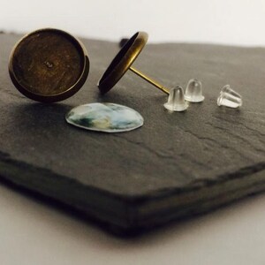 May include: Two gold-toned earring settings with a clear glass cabochon and three clear silicone earring backs on a black slate surface.
