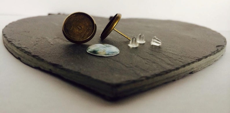 May include: Two gold-toned earring settings with a clear glass cabochon and three clear silicone earring backs on a black slate surface.