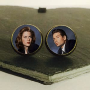 May include: A pair of round, gold-toned earrings featuring images of a woman and a man. The woman has brown hair and is wearing a blue jacket. The man has brown hair and is wearing a blue suit jacket.