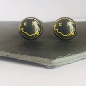 Puede incluir: Un par de pendientes de botón negros con un diseño de cara sonriente amarilla. La cara sonriente tiene una 'X' para los ojos y una lengua que sobresale.