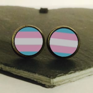 May include: A pair of round earrings with a transgender pride flag design. The earrings are on a dark gray slate surface.
