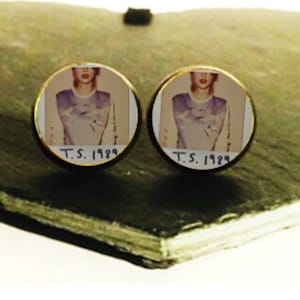 May include: A pair of gold cufflinks with a round, white background featuring a vintage photograph of a person wearing a light blue shirt. The text "T.S. 1929" is printed on the bottom of each cufflink.