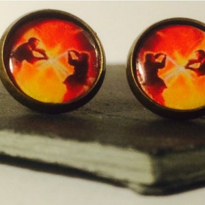 May include: A pair of round, bronze-colored earrings with a red, orange, and yellow image of two figures fighting with lightsabers.