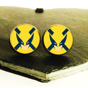 May include: Two circular stud earrings with a yellow base and a blue and white graphic. They are presented on a heart-shaped, dark grey stone. The earrings are a stylish accessory.