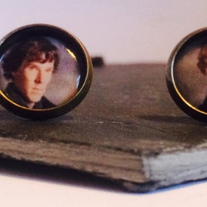 May include: Two round, bronze-colored cufflinks with images of men's faces. The cufflinks are on a dark gray surface.