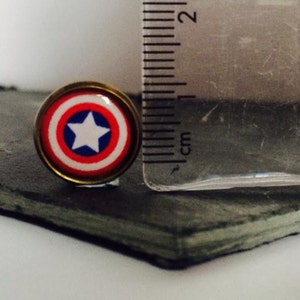 May include: A close-up of a small, round, red, white, and blue Captain America shield pin. The pin is about 1 cm in diameter and is on a black slate surface.