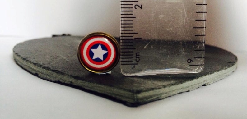May include: A close-up of a small, round, red, white, and blue Captain America shield pin. The pin is about 1 cm in diameter and is on a black slate surface.