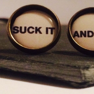 Puede incluir: Dos pequeños pendientes redondos con un acabado bronce. Los pendientes tienen un fondo blanco con texto negro. El pendiente izquierdo dice "SUCK IT" y el pendiente derecho dice "AND SEE".