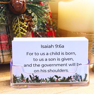 May include: A clear acrylic card holder with a stack of white cards. The cards display the text "Isaiah 9:6a" and a religious quote. The holder is decorated with a festive green and red floral design, with a blurred background of Christmas decorations.
