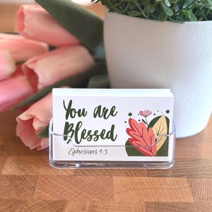 May include: A clear acrylic card holder displaying a stack of white cards. The cards feature the text "You are Blessed" in green script, with "Ephesians 1:3" below. A floral design with red, pink, and green leaves is on the right.
