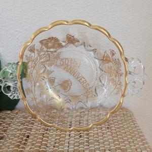 May include: Clear glass serving bowl with a gold rim and handles. The bowl features gold floral designs and a banner that reads "50th Anniversary". A decorative piece for special occasions.