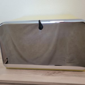 May include: A rectangular bread box with a pale yellow body and a large, reflective chrome-finished front door. The door features a single, small, black teardrop-shaped handle centered at the top edge for opening the compartment.