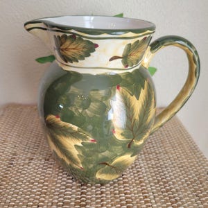 May include: A green ceramic pitcher with a white rim and a handle. The pitcher is decorated with a leafy pattern in shades of green, yellow, and red.