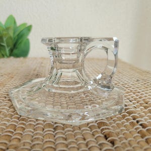 May include: A clear glass candlestick holder with a square base and a circular handle. The candlestick holder has a detailed, textured design and is made of transparent glass. The base is hexagonal in shape.