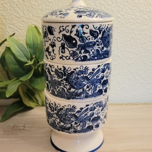 May include: A three-tiered ceramic stacking jar with a lid, featuring a white base and intricate blue floral and bird patterns. The cylindrical container rests on a pedestal base and is topped with a pointed finial handle on the lid.