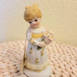 May include: A porcelain figurine of a young girl with blonde hair, wearing a white dress with yellow accents and floral patterns. She holds a bouquet of white flowers. The figurine has a delicate, vintage aesthetic.
