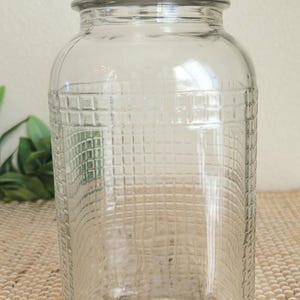 May include: A clear glass jar with a textured grid pattern and a metal lid. The jar has a wide body and a slightly narrower neck. The lid is a dark gray color. The jar is sitting on a woven surface.