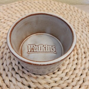 May include: A small, round, ceramic bowl with a light beige exterior and a darker brown rim. The word "Watkins" is embossed in the center of the bowl. The bowl is sitting on a woven, beige placemat.